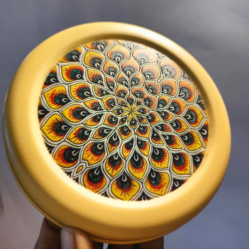 A yellow metal tiffin lid with a colorful peacock feather mandala design.