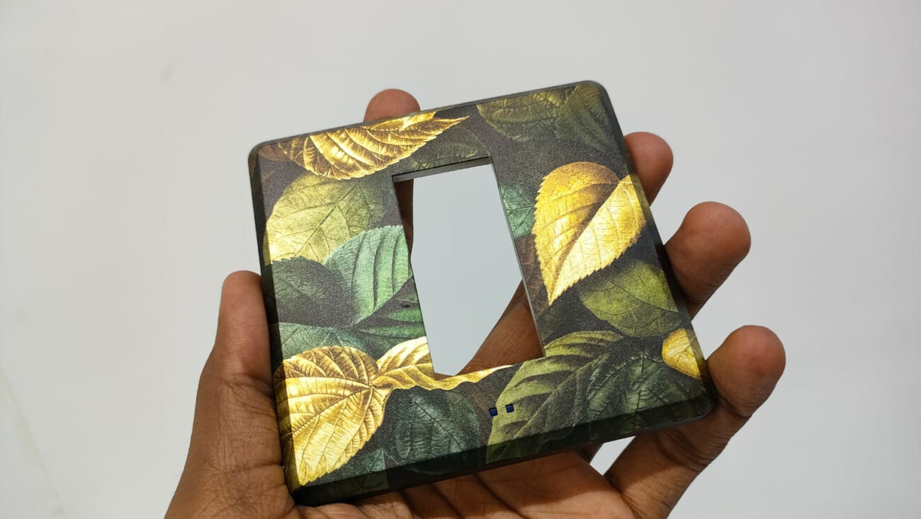 A hand holding a rectangular switchboard cover with a vibrant leaf pattern in shades of green and gold.