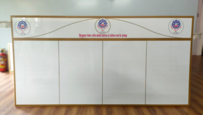 A rectangular acrylic signage board with four blank white sections and a curved banner with logos and text in Hindi.