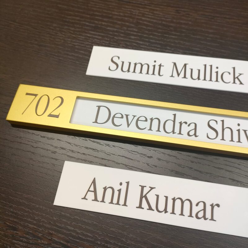 A gold-colored acrylic name plate with the number 702, the names Devendra Shiv and Anil Kumar, and two additional name cards.